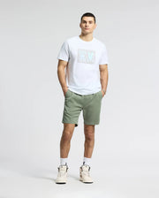 A man stands against a plain background wearing a white Psycho Bunny MENS GOLD COAST GRAPHIC TEE - B6U631F200, green shorts, and white sneakers, with his hands in his pockets and short hair.