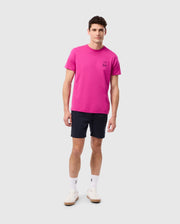 A man stands against a plain white background. He is wearing a bright pink MENS PEARLAND BACK GRAPHIC TEE - B6U632C200 by Psycho Bunny, featuring an embroidered bunny logo on the left chest, paired with dark blue shorts. He has white socks and white sneakers. He is looking straight ahead with a neutral expression.