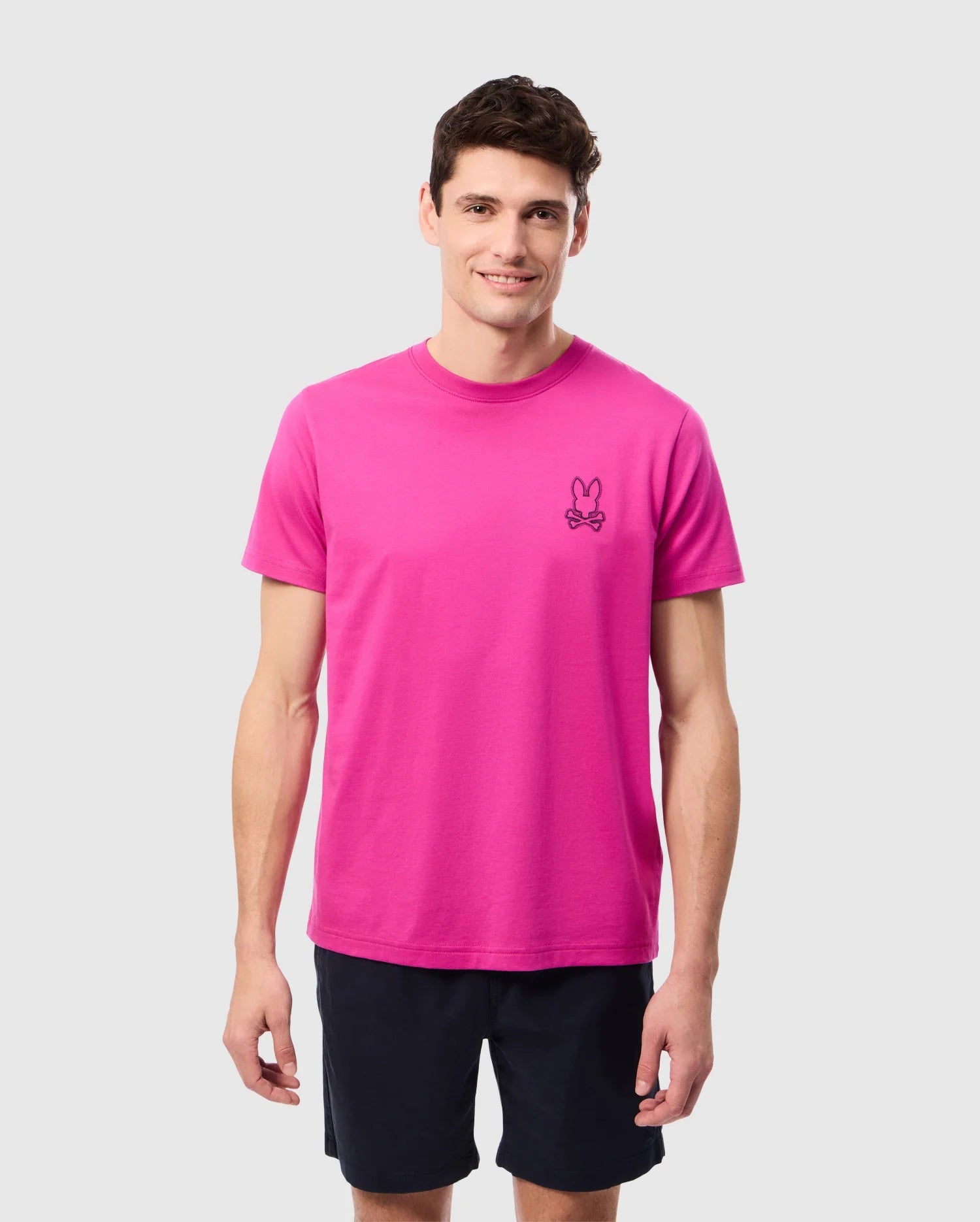 A man stands against a plain background wearing the MENS PEARLAND BACK GRAPHIC TEE - B6U632C200 from Psycho Bunny in vibrant pink, featuring a small embroidered bunny logo on the left side of his chest. Made from luxurious Pima cotton, the shirt perfectly complements his short dark hair and cheerful smile. He is also dressed in dark shorts.