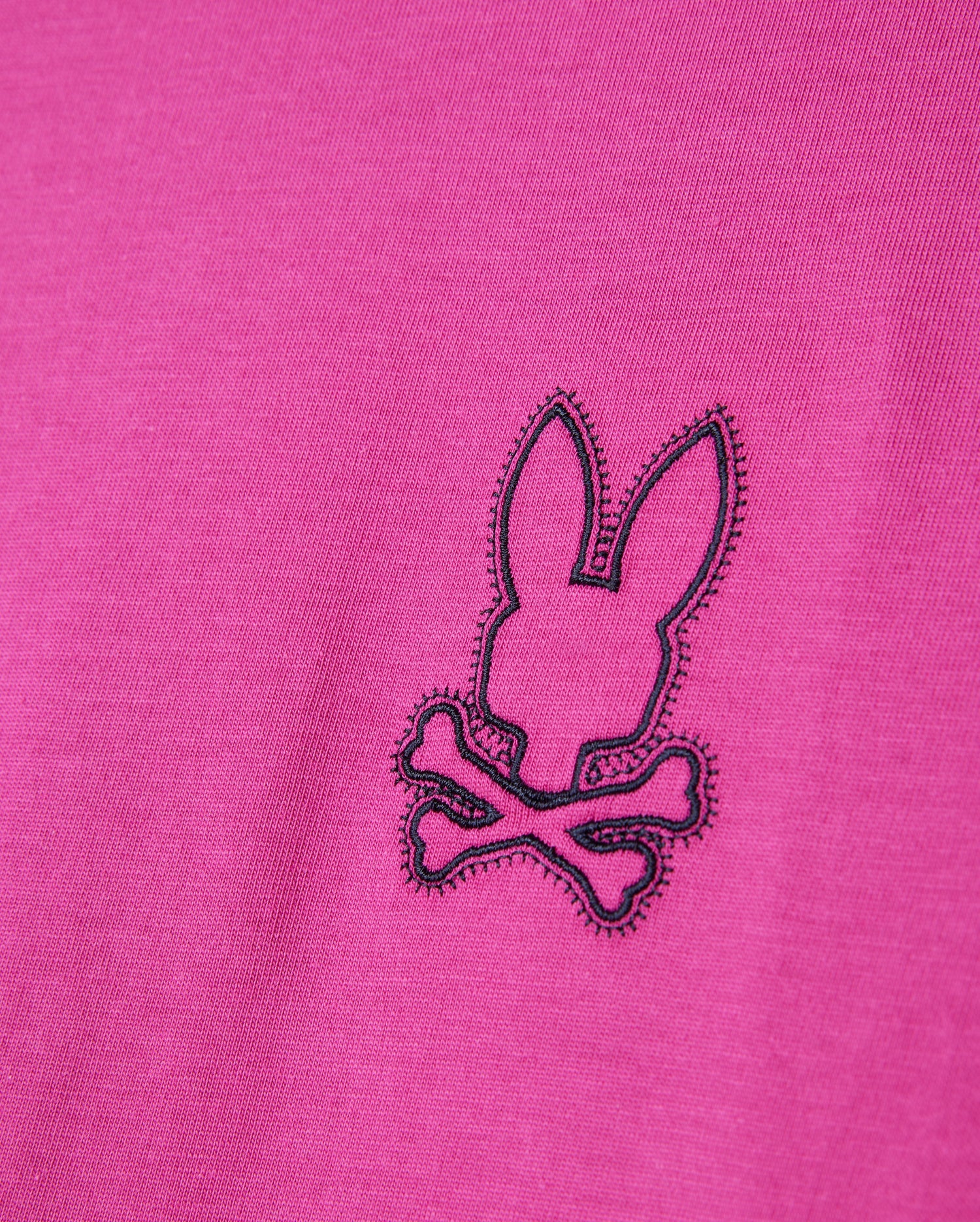 Close-up of the Psycho Bunny MEN'S PEARLAND BACK GRAPHIC TEE - B6U632C200 in pink, showcasing an embroidered black outline of a bunny head above two crossed bones, creating a playful pirate-inspired logo. The stitching is intricate, offering a textured contrast to the Pima cotton fabric.