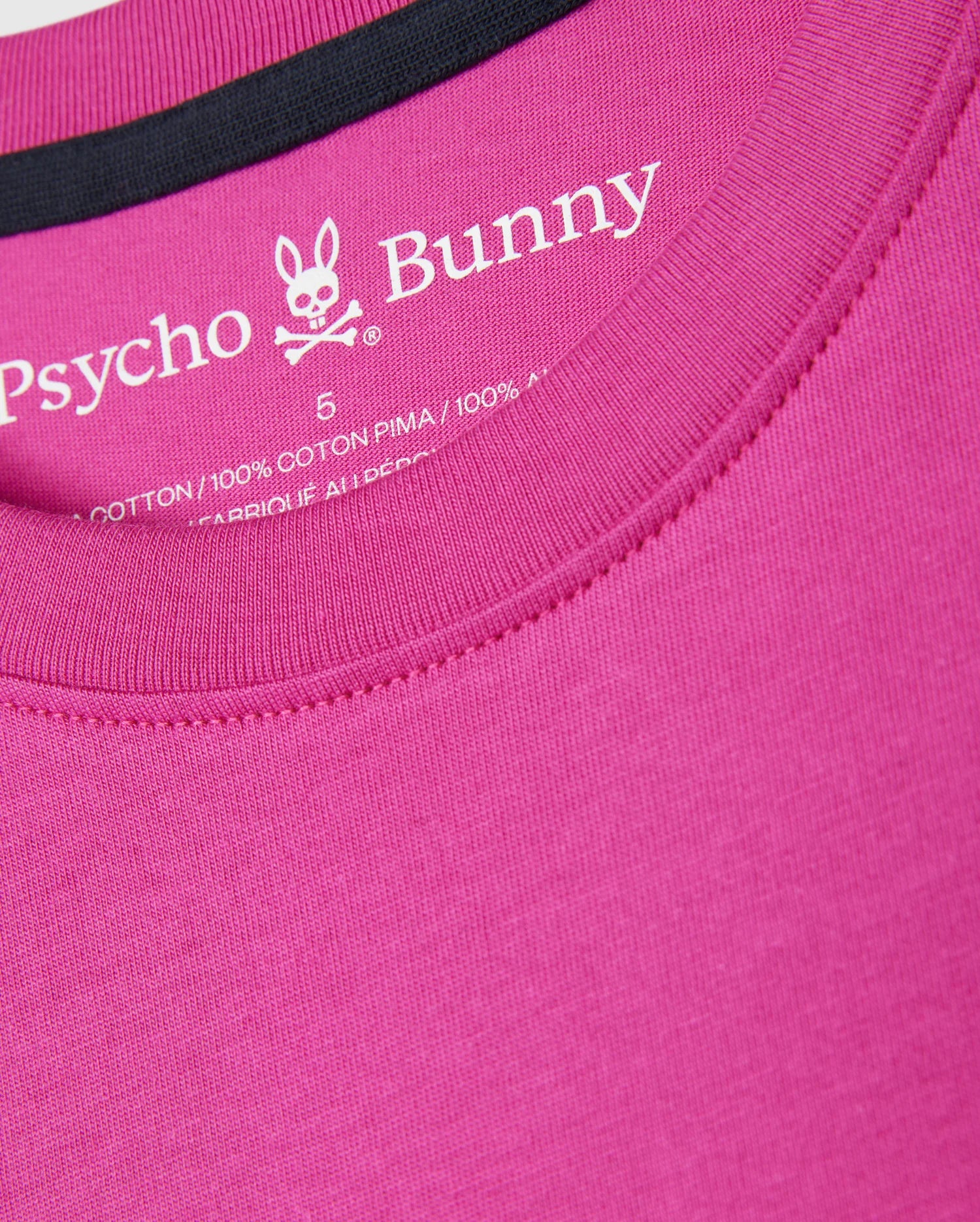 Close-up of a pink Psycho Bunny Mens Pearland Back Graphic Tee (B6U632C200) with an embroidered white label inside the collar. The label features the Psycho Bunny logo, a bunny skull with crossbones, and material information, indicating the shirt is made of 100% Pima cotton.