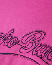 A close-up of the Psycho Bunny MENS PEARLAND BACK GRAPHIC TEE - B6U632C200 in soft pink Pima cotton, showcasing intricate black embroidered cursive letters. The bold and heavy embroidery contrasts sharply against the vivid pink background, with detailed stitching and fabric texture clearly visible.