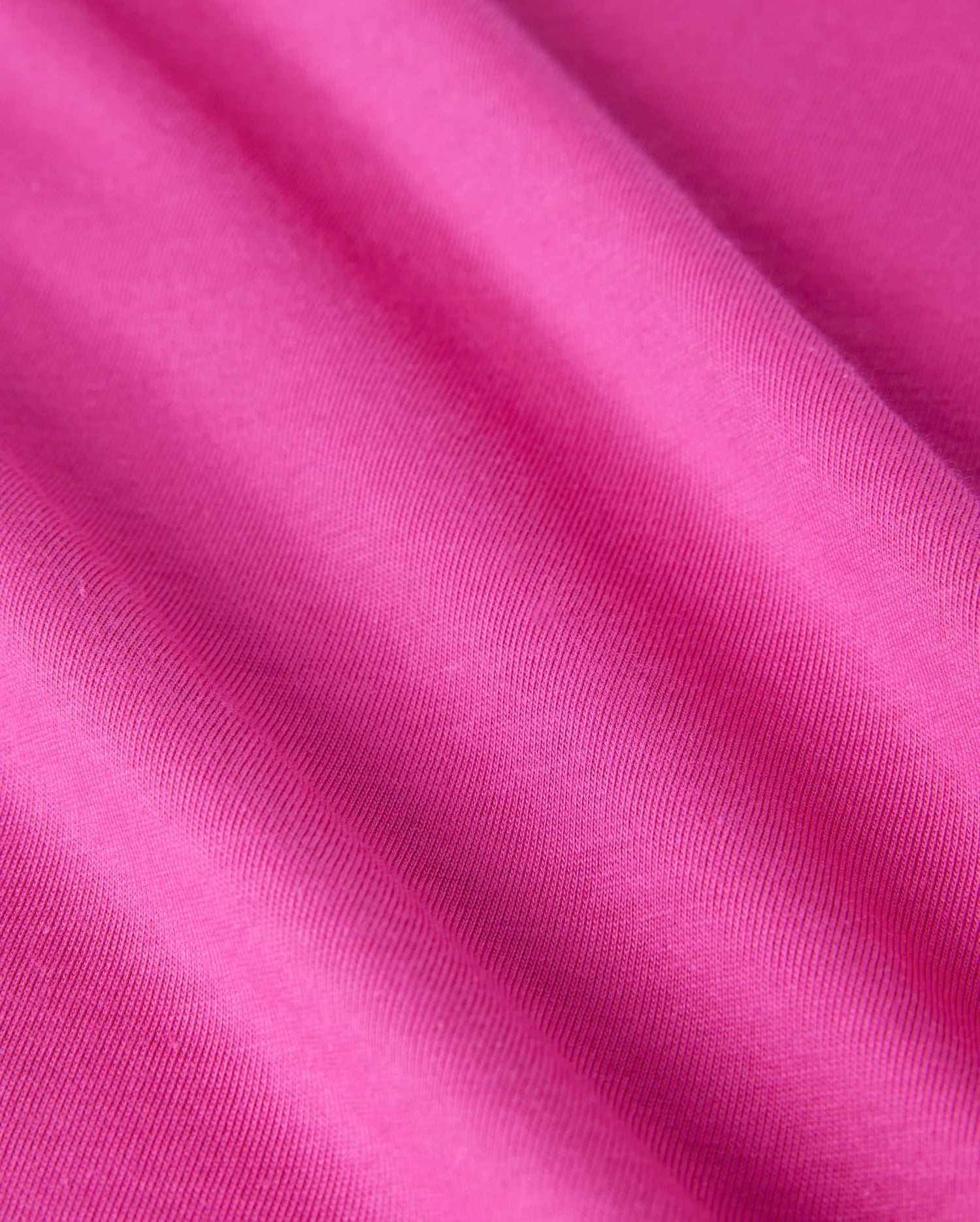 Close-up shot of a pink Men's Pearland Back Graphic Tee (B6U632C200) by Psycho Bunny, showcasing its smooth Pima cotton texture and slight folds. The material appears to be soft and uniform in color, with visible weave patterns in the textile and a delicate embroidered detail.