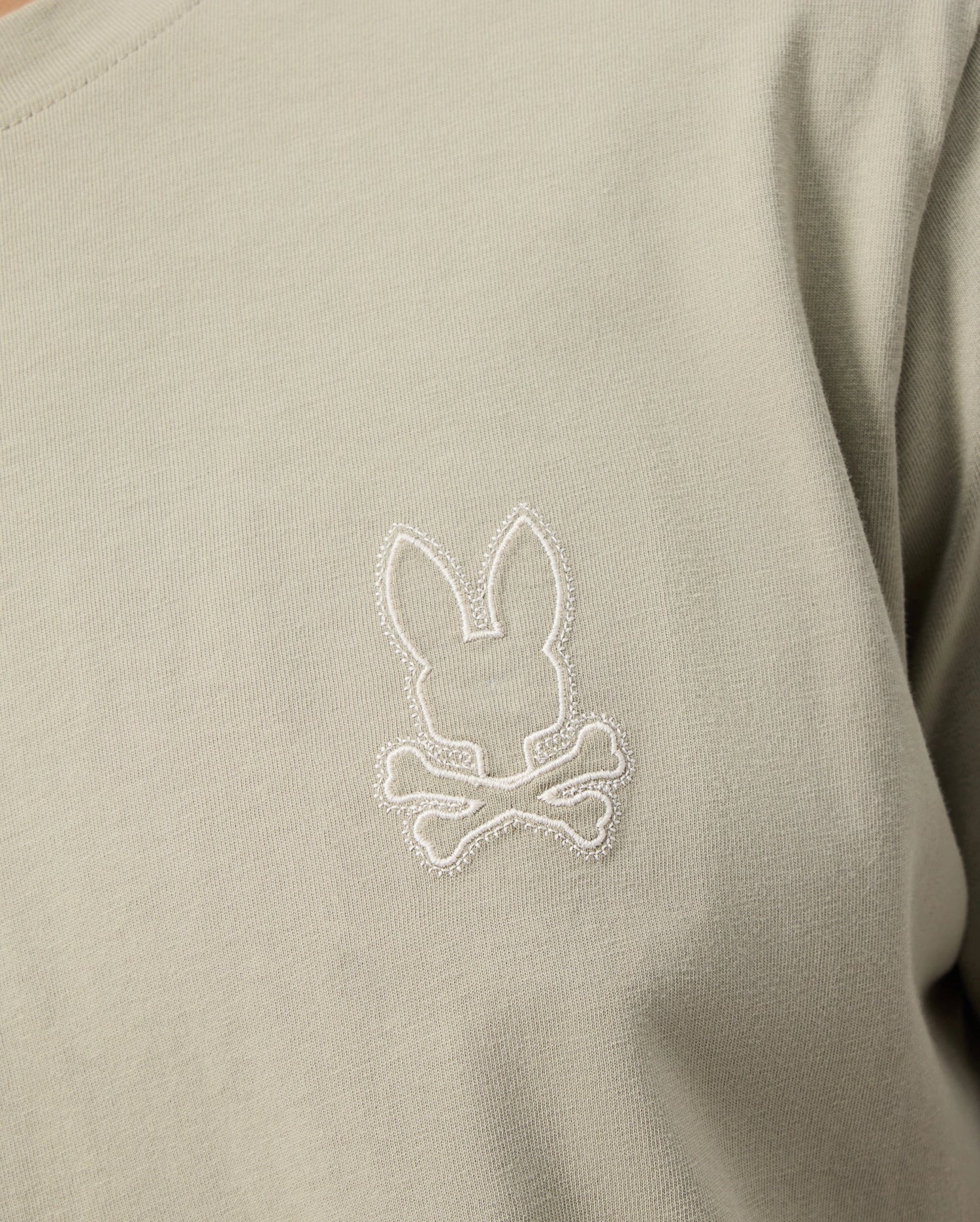 A close-up image of the Psycho Bunny MENS PEARLAND BACK GRAPHIC TEE - B6U632C200 in light grey-green, featuring an embroidered rabbit head above a pair of crossed bones on the chest. The intricate stitching is done in white, creating a chic contrast with the Pima cotton fabric.