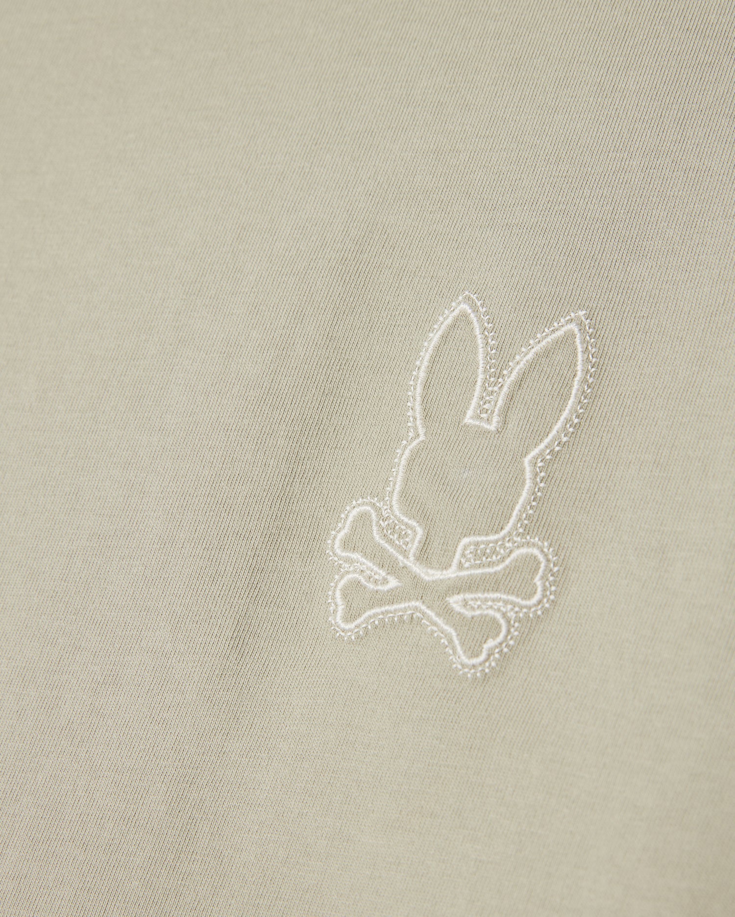 A close-up of light beige Pima cotton fabric from the Psycho Bunny MENS PEARLAND BACK GRAPHIC TEE - B6U632C200, featuring an embroidered design of a bunny head above crossed bones, reminiscent of a skull and crossbones motif. The embroidery is white, creating a subtle contrast against the high-quality Pearland tee.