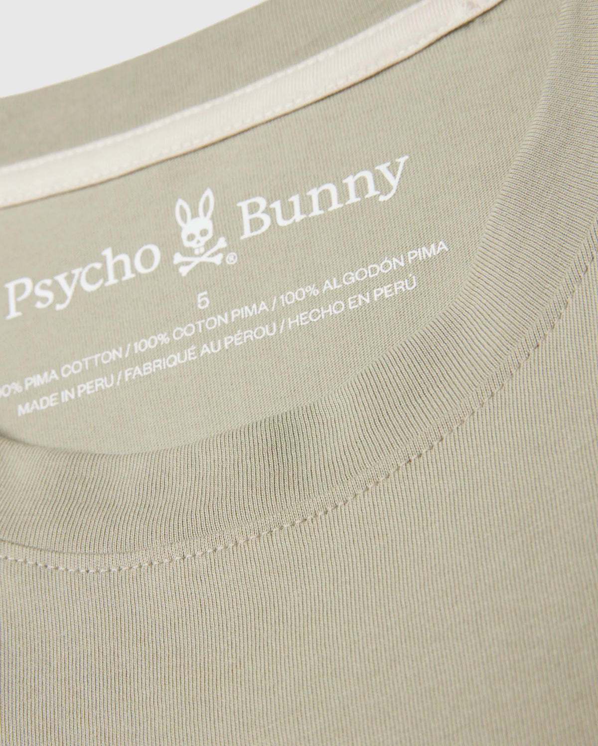 Close-up of the neckline of a beige MENS PEARLAND BACK GRAPHIC TEE - B6U632C200 showcasing the embroidered brand name "Psycho Bunny" beneath a bunny logo. The text includes "100% Pima Cotton," size "5," and "Made in Peru" written in English, French, and Spanish. The fabric features a ribbed texture.
