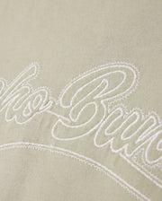 A close-up of the light Pima cotton fabric on the MENS PEARLAND BACK GRAPHIC TEE - B6U632C200 by Psycho Bunny, showcasing the white embroidered text "Club Bure" in a cursive font. The embroidery is slightly raised above the fabric surface, creating a textured effect.