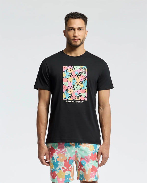 A man wears the Psycho Bunny MENS LEMMON GRAPHIC TEE in black Pima cotton, featuring a vibrant abstract floral rectangle. His shorts complement the tee with a similar floral pattern on a light background, set against a plain white backdrop that highlights the vibrant graphics on his chest.