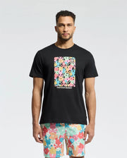 A man wears the Psycho Bunny MENS LEMMON GRAPHIC TEE in black Pima cotton, featuring a vibrant abstract floral rectangle. His shorts complement the tee with a similar floral pattern on a light background, set against a plain white backdrop that highlights the vibrant graphics on his chest.