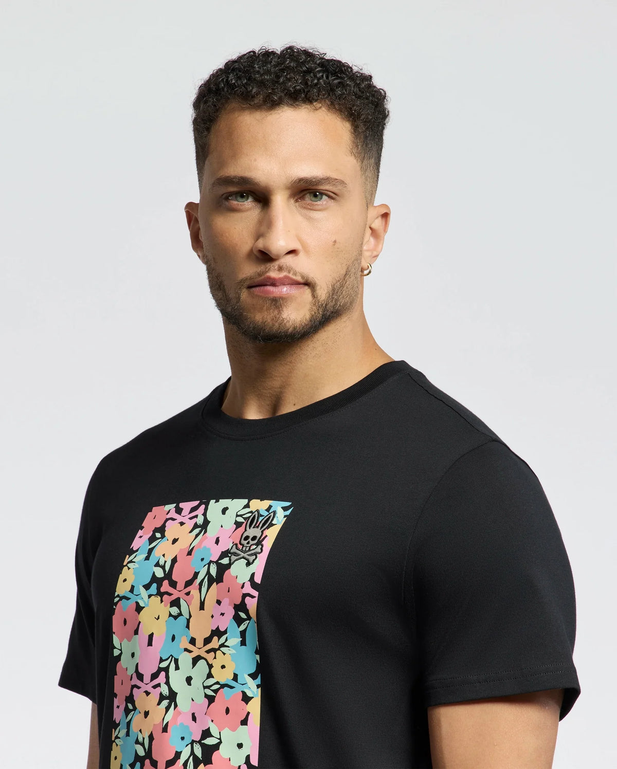 A man with short curly hair and a beard wears the MENS LEMMON GRAPHIC TEE by Psycho Bunny featuring a vibrant floral design and subtle bunny graphic, ensuring all-day comfort with its soft Pima cotton fabric as he stands against a plain white background.