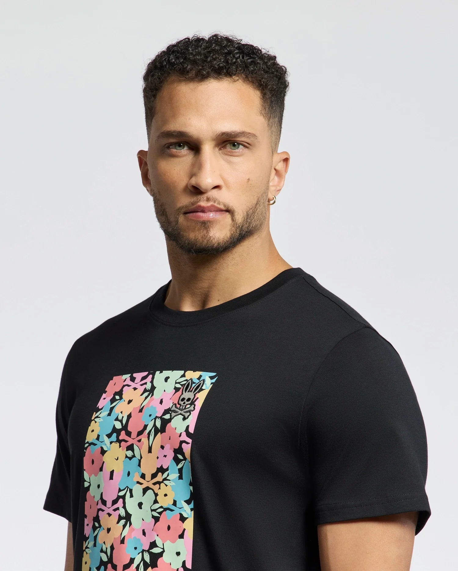 A man with short curly hair and a beard wears the MENS LEMMON GRAPHIC TEE by Psycho Bunny featuring a vibrant floral design and subtle bunny graphic, ensuring all-day comfort with its soft Pima cotton fabric as he stands against a plain white background.