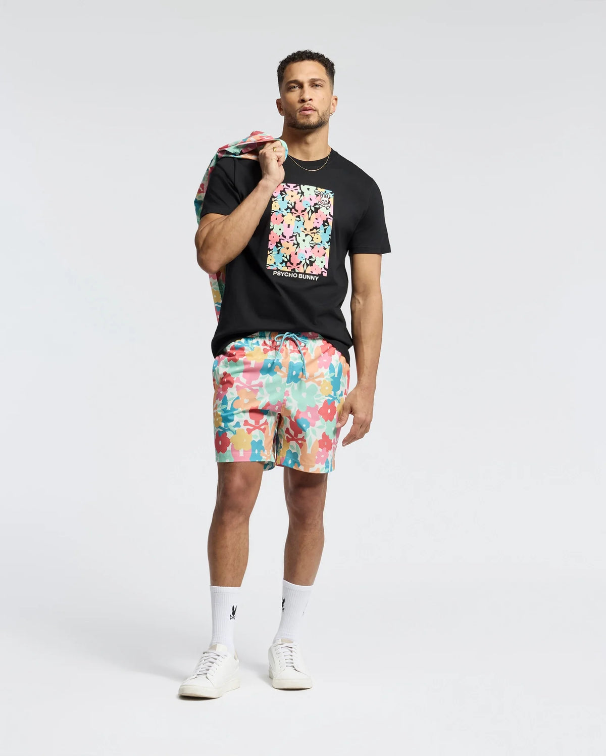 A person exudes confidence in a Psycho Bunny MENS LEMMON GRAPHIC TEE - B6U655F200 made of black Pima cotton with a colorful bunny graphic. They pair it with vibrant shorts and white sneakers, draping a matching patterned jacket over one shoulder against the plain light background.