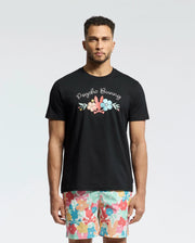 A man in a black Psycho Bunny MENS MEYLON GRAPHIC TEE - B6U656F200 with vibrant floral and text designs stands against a plain light background. His multicolored floral-patterned shorts enhance the casual flair of this Pima cotton ensemble, perfect for a sunny day out.