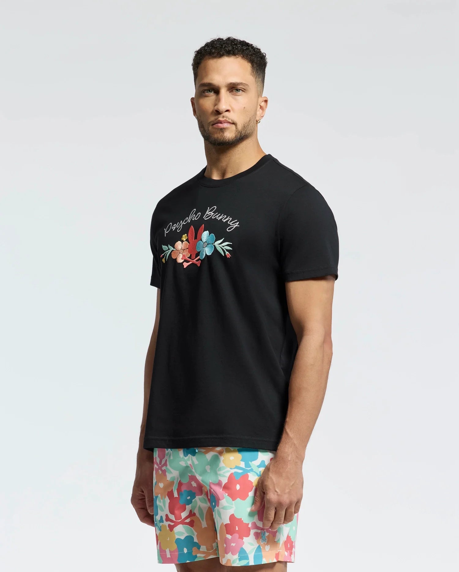 A man with short curly hair and a beard stands against a neutral background in a Psycho Bunny B6U656F200 Pima cotton T-shirt with colorful floral design and text, paired with multi-colored floral shorts, gazing into the camera.