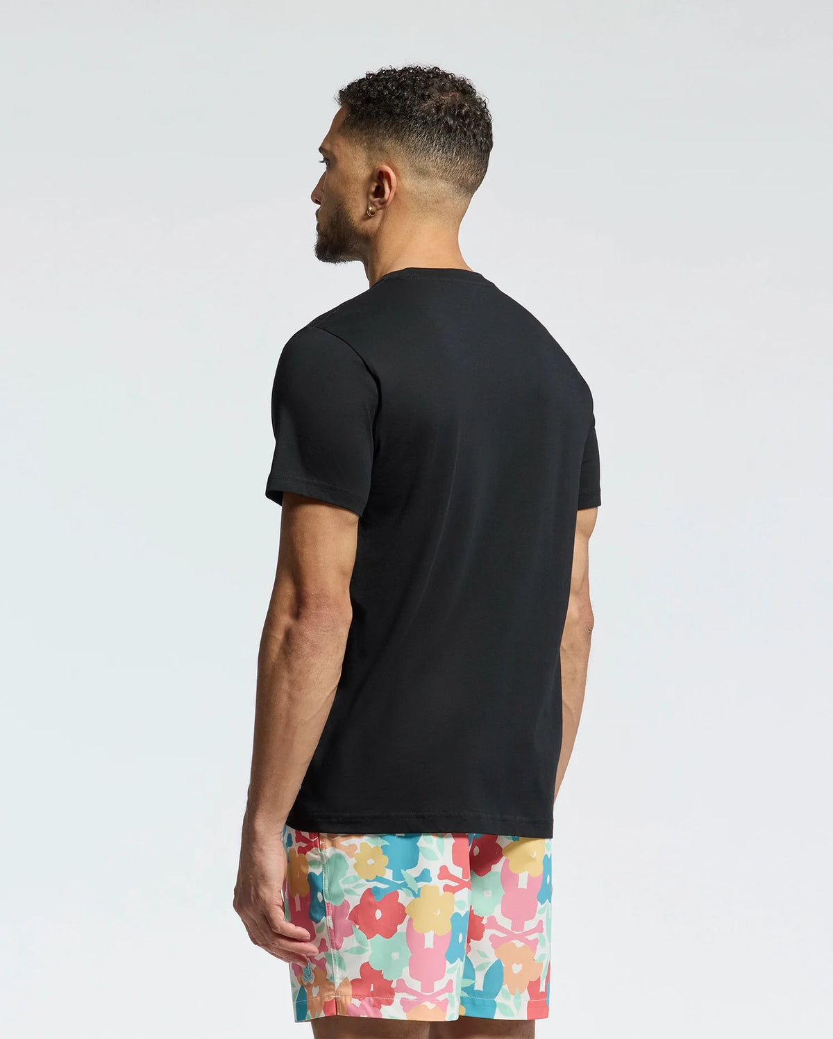 A man with short, curly hair stands with his back to the camera, wearing a Psycho Bunny MENS MEYLON GRAPHIC TEE (B6U656F200) and colorful shorts with an abstract floral design. The background is a simple light gradient.