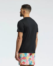 A man with short, curly hair stands with his back to the camera, wearing a Psycho Bunny MENS MEYLON GRAPHIC TEE (B6U656F200) and colorful shorts with an abstract floral design. The background is a simple light gradient.