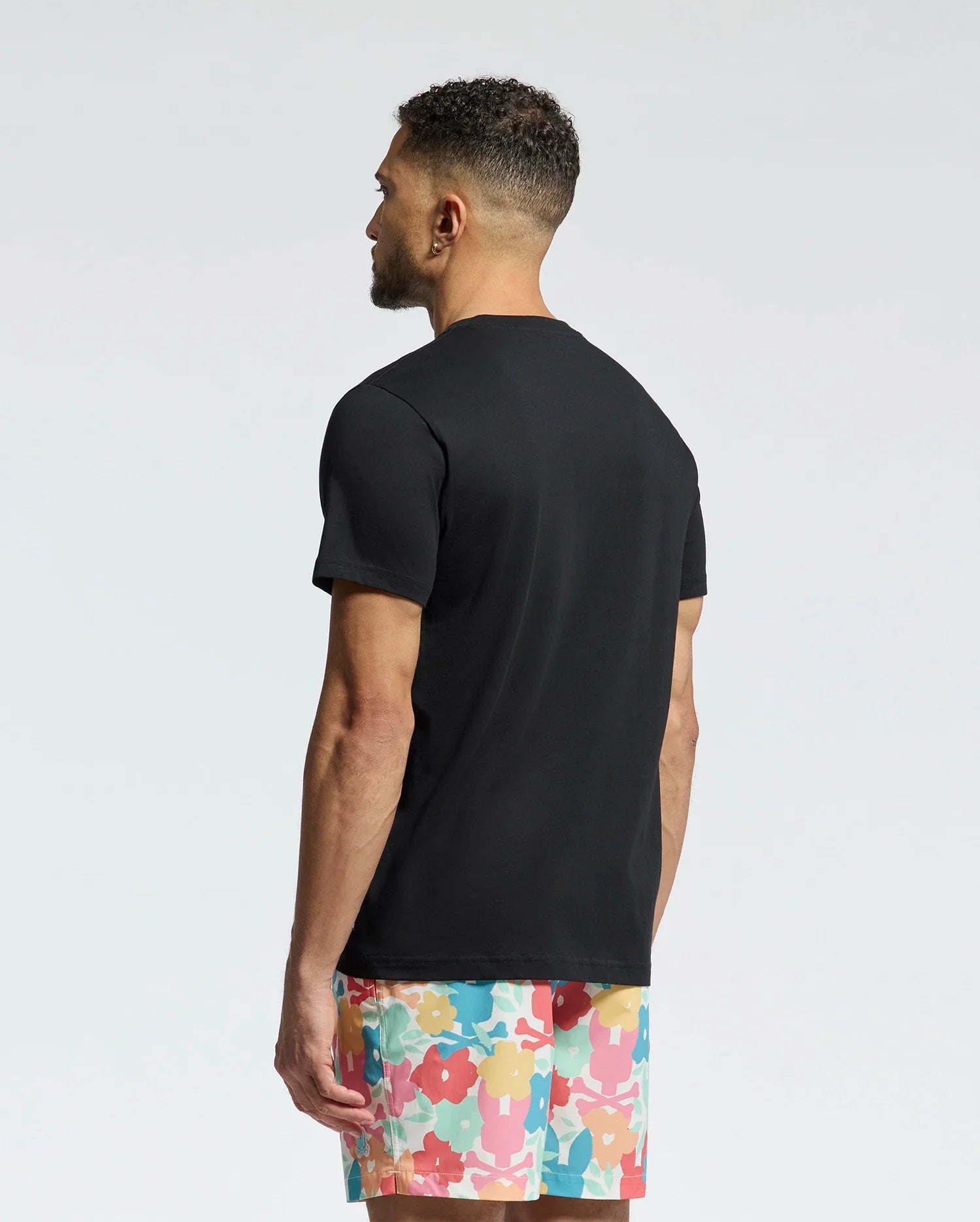 A man with short, curly hair stands with his back to the camera, wearing a Psycho Bunny MENS MEYLON GRAPHIC TEE (B6U656F200) and colorful shorts with an abstract floral design. The background is a simple light gradient.