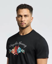 A curly-haired, bearded man wears a black MENS MEYLON GRAPHIC TEE - B6U656F200 by Psycho Bunny, crafted from soft Pima cotton with floral embroidery and "Psycho Bunny" text against a light-colored plain background.