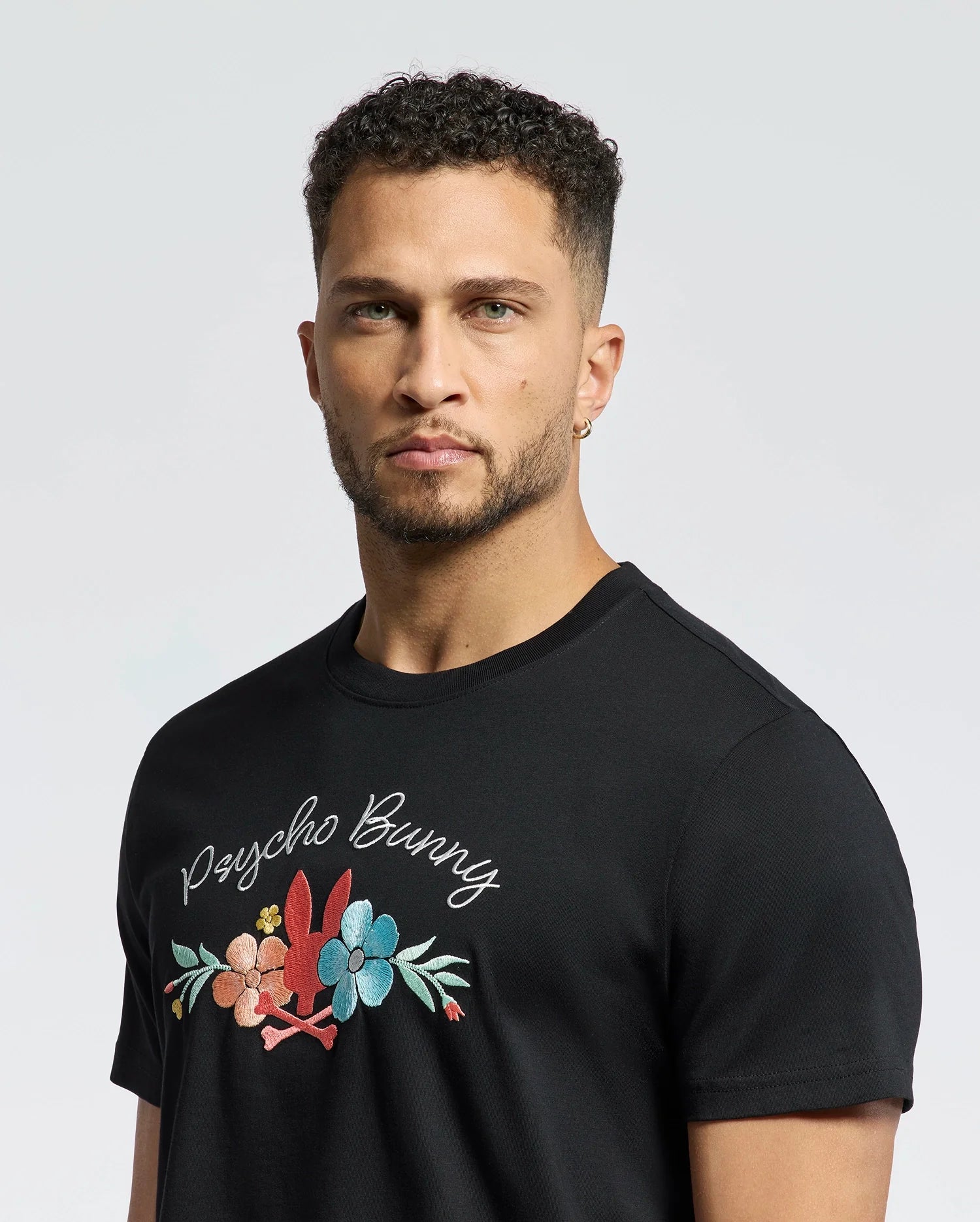A curly-haired, bearded man wears a black MENS MEYLON GRAPHIC TEE - B6U656F200 by Psycho Bunny, crafted from soft Pima cotton with floral embroidery and "Psycho Bunny" text against a light-colored plain background.