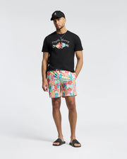 A man stands against a plain background wearing a black baseball cap, a Psycho Bunny MENS MEYLON GRAPHIC TEE - B6U656F200 with floral graphics, colorful floral-patterned shorts, and black slides.