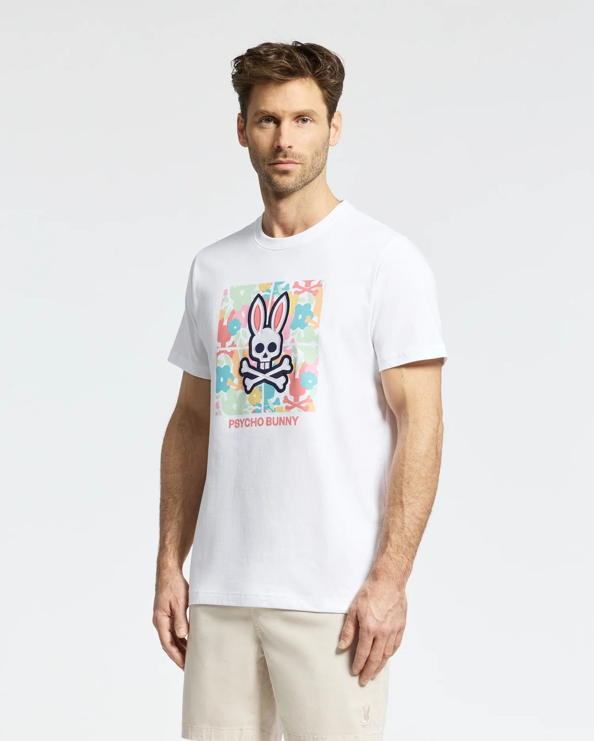 A man with short hair stands against a plain background wearing the MENS LEMORA GRAPHIC TEE - B6U657F200 by Psycho Bunny, a white Pima cotton T-shirt with a vibrant water-based bunny and crossbones print. He pairs it with light-colored shorts.
