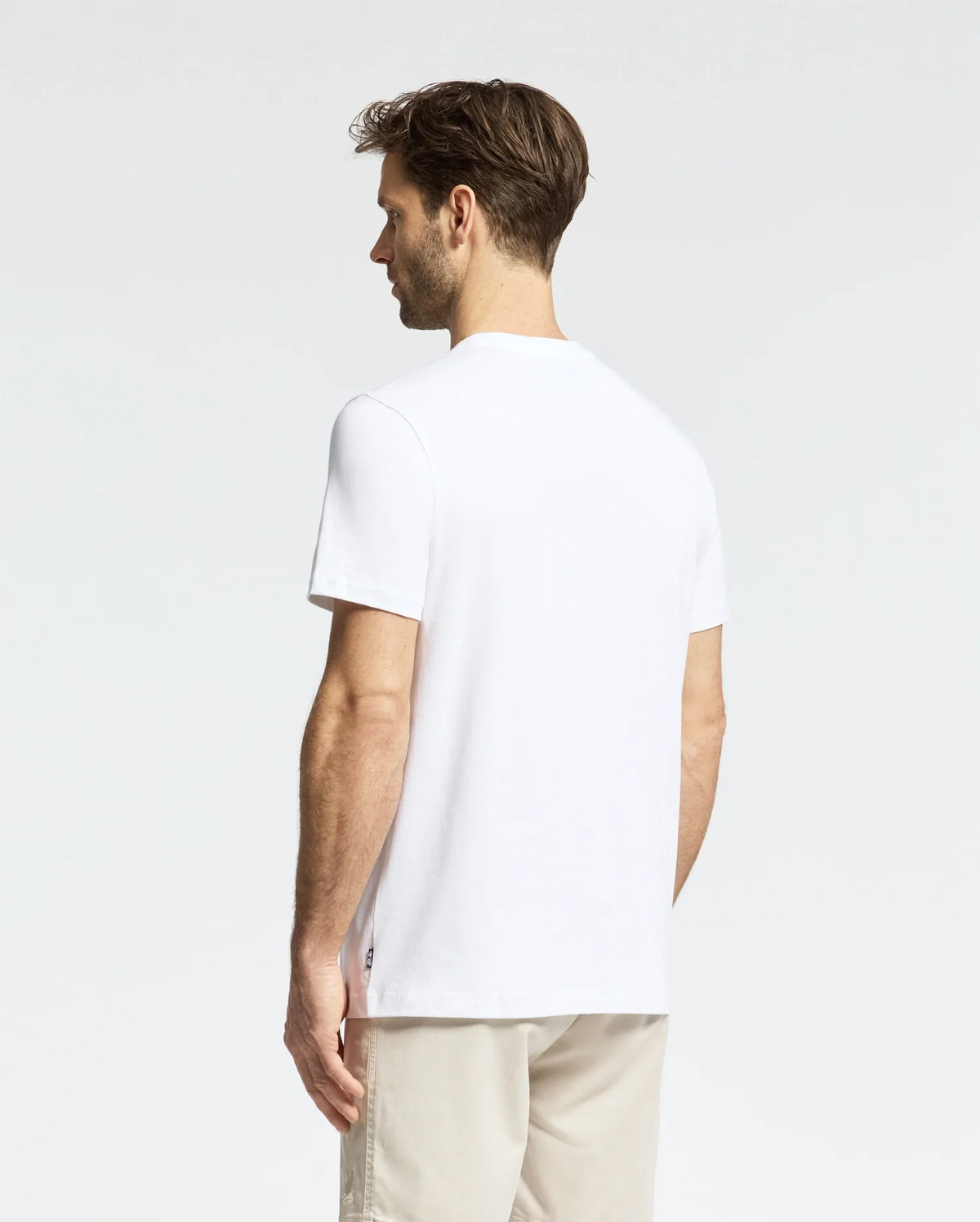 A man with short brown hair stands facing away, donning a MENS LEMORA GRAPHIC TEE - B6U657F200 by Psycho Bunny and beige pants. He is set against a plain white backdrop, exuding a relaxed posture.