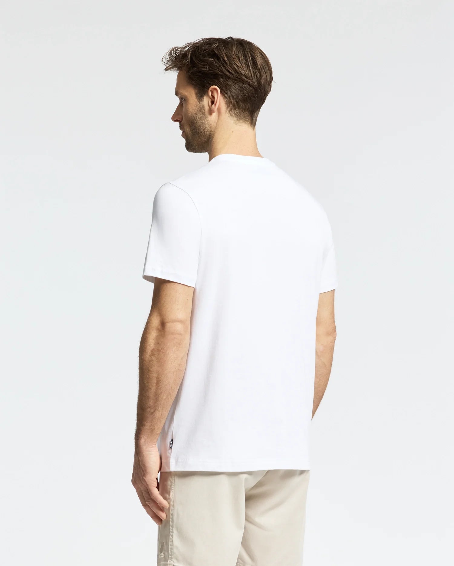 A man with short brown hair stands facing away, donning a MENS LEMORA GRAPHIC TEE - B6U657F200 by Psycho Bunny and beige pants. He is set against a plain white backdrop, exuding a relaxed posture.