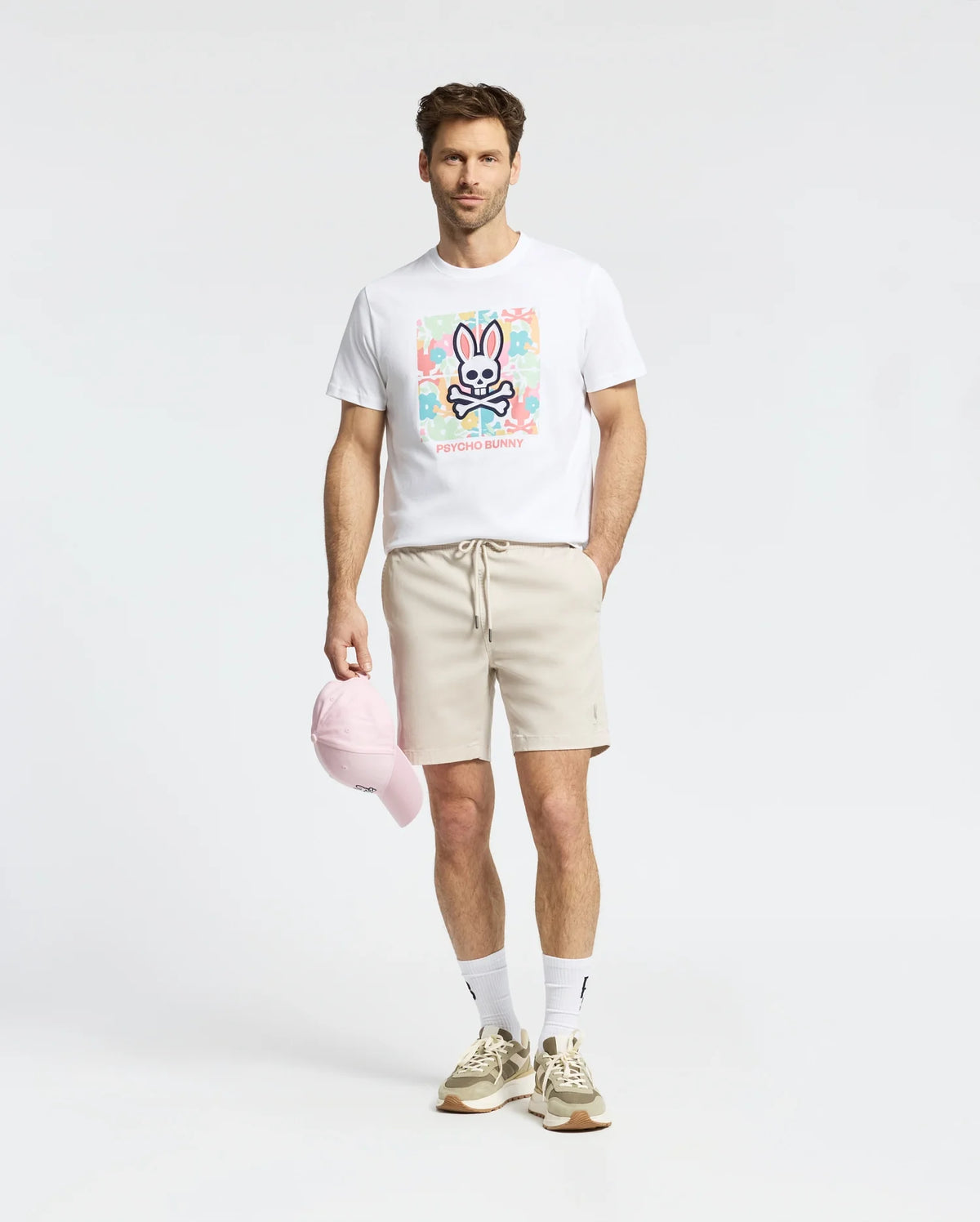 A man in a white Mens Lemora Graphic Tee by Psycho Bunny with a colorful print logo, beige shorts, white socks, beige sneakers, and holding a pink cap stands against a light gray background.