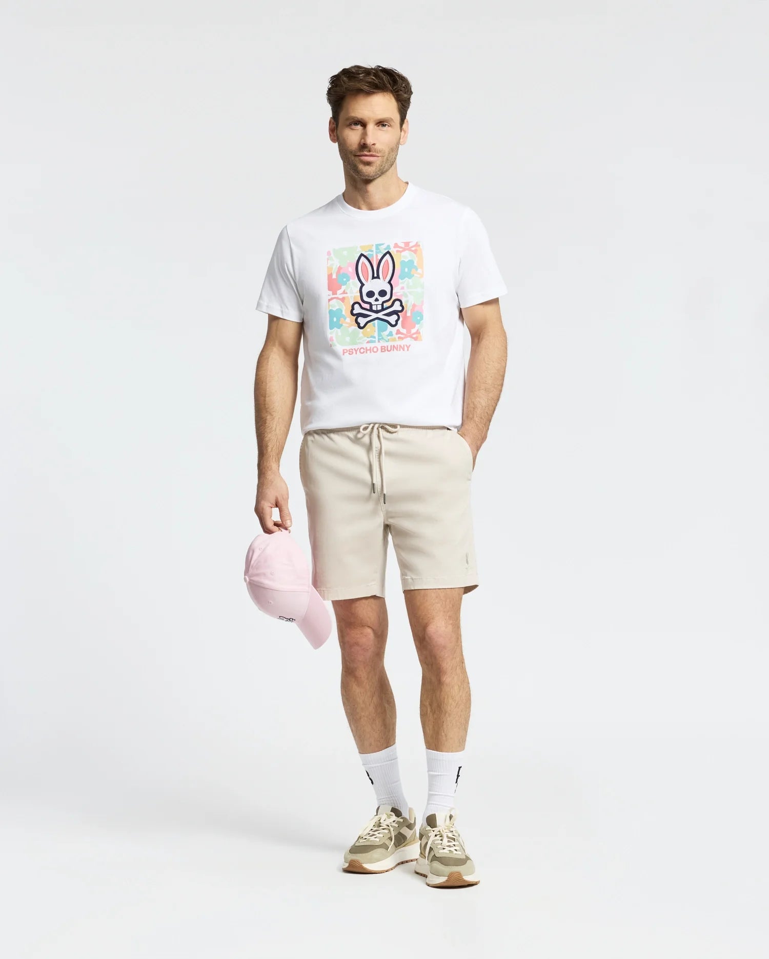 A man in a white Mens Lemora Graphic Tee by Psycho Bunny with a colorful print logo, beige shorts, white socks, beige sneakers, and holding a pink cap stands against a light gray background.