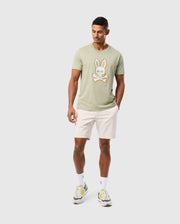 A man with short dark hair is wearing a light green **MENS KAYDEN GRAPHIC TEE - B6U676C200** featuring an embroidered bunny and crossbones motif by **Psycho Bunny**, paired with light beige shorts. He also has on white socks and green, white, and yellow sneakers. He is standing against a plain white background, looking downward.