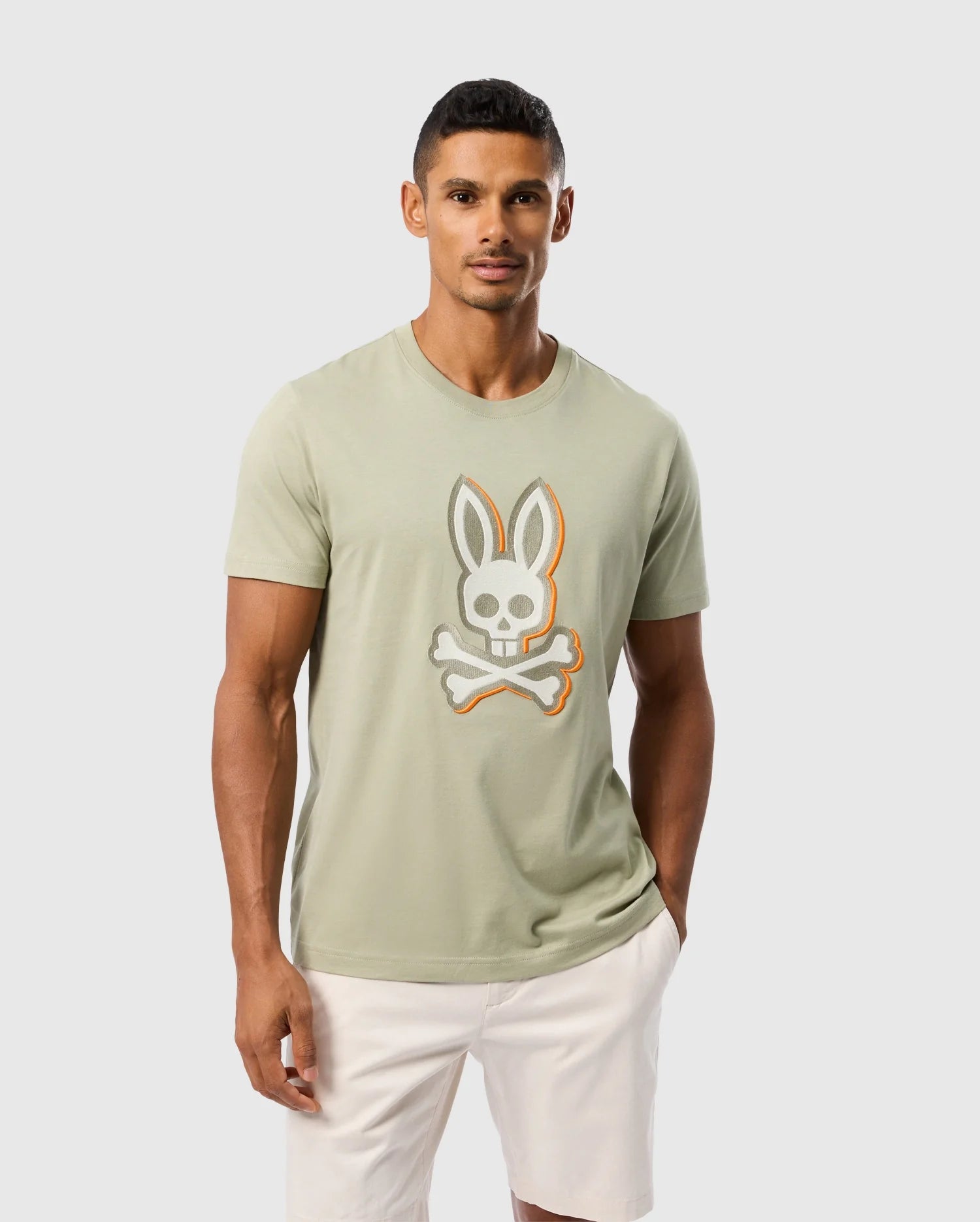 A man stands against a plain white background, wearing a light green MENS KAYDEN GRAPHIC TEE - B6U676C200 by Psycho Bunny featuring an embroidered bunny skull and crossbones logo, with white shorts. His hands are in his pockets, and he gazes directly at the camera.