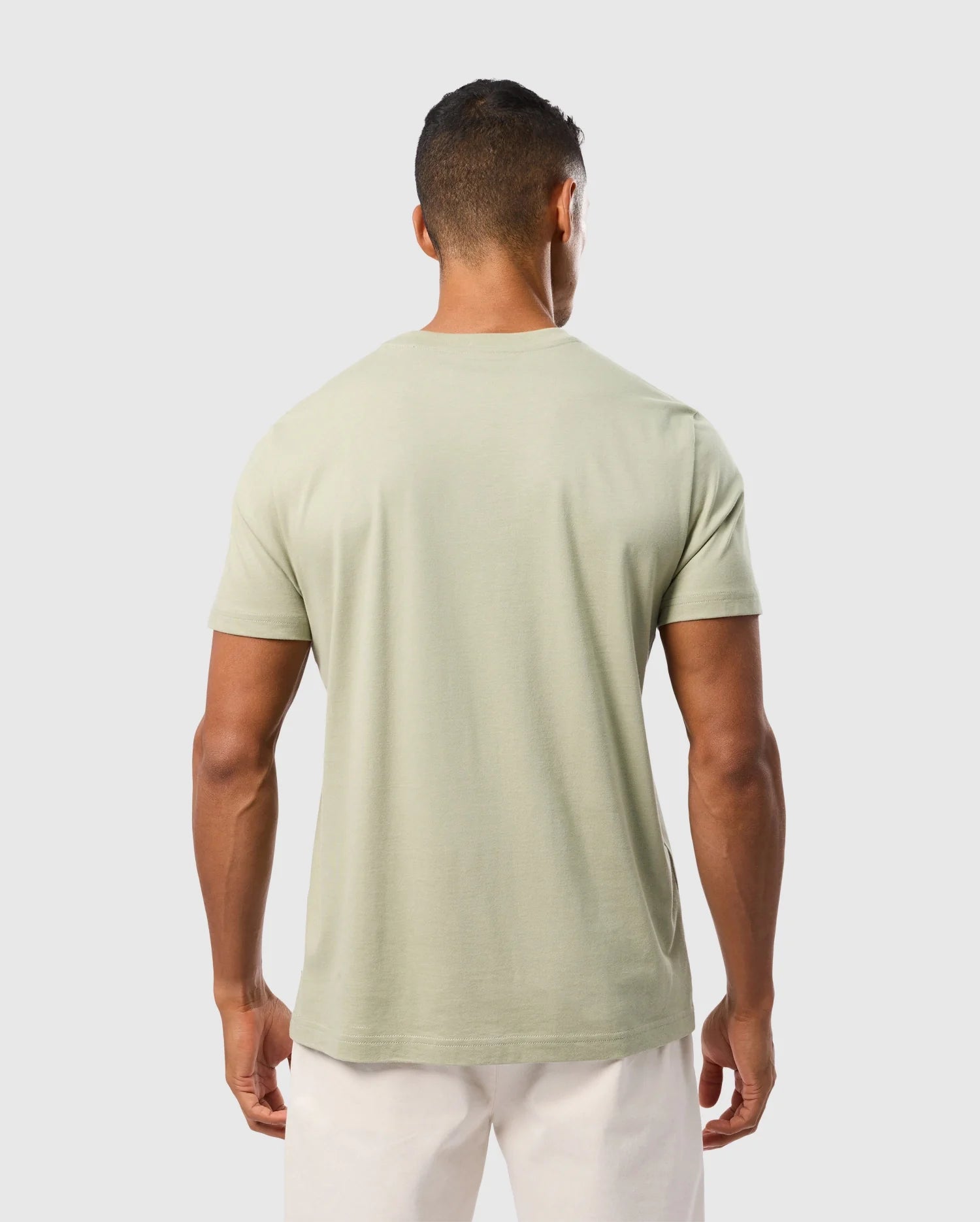 A man is standing with his back to the camera, wearing a light green Psycho Bunny MENS KAYDEN GRAPHIC TEE - B6U676C200 and white pants. The background is plain and light-colored. The man has short hair and an athletic build.