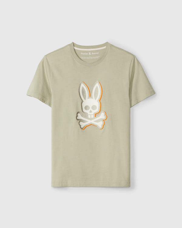 A light beige MENS KAYDEN GRAPHIC TEE - B6U676C200 featuring a stylized design of a skull with bunny ears and crossbones on the front center. Made from soft Pima cotton, the t-shirt displays the brand label "Psycho Bunny" inside the collar. The t-shirt is showcased against a plain white background.