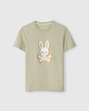 A light beige MENS KAYDEN GRAPHIC TEE - B6U676C200 featuring a stylized design of a skull with bunny ears and crossbones on the front center. Made from soft Pima cotton, the t-shirt displays the brand label "Psycho Bunny" inside the collar. The t-shirt is showcased against a plain white background.