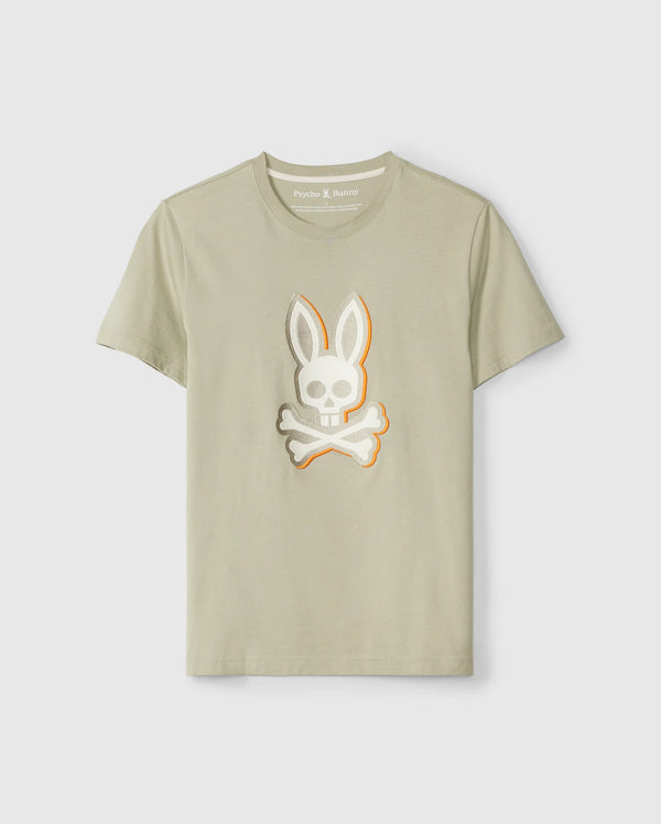 A light beige MENS KAYDEN GRAPHIC TEE - B6U676C200 featuring a stylized design of a skull with bunny ears and crossbones on the front center. Made from soft Pima cotton, the t-shirt displays the brand label "Psycho Bunny" inside the collar. The t-shirt is showcased against a plain white background.