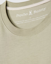 Close-up image of a light green MENS KAYDEN GRAPHIC TEE - B6U676C200. The label inside the neckline reads "Psycho Bunny," followed by an embroidered bunny logo and details about the material, indicating it is 100% Pima cotton and made in Peru.