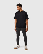 A man wearing a Psycho Bunny MENS DUKE OVERSIZED TEE - B6U742D200 in heavyweight Pima cotton, dark pants, and white sneakers stands against a plain light gray background. He looks down to his left with hands relaxed at his sides. His outfit is casual and minimalistic.