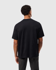 A person with short curly hair is standing and facing away from the camera, showcasing the relaxed fit of the Psycho Bunny MENS DUKE OVERSIZED TEE (B6U742D200) in plain black heavyweight cotton, paired with dark gray pants. The background is plain and light-colored.