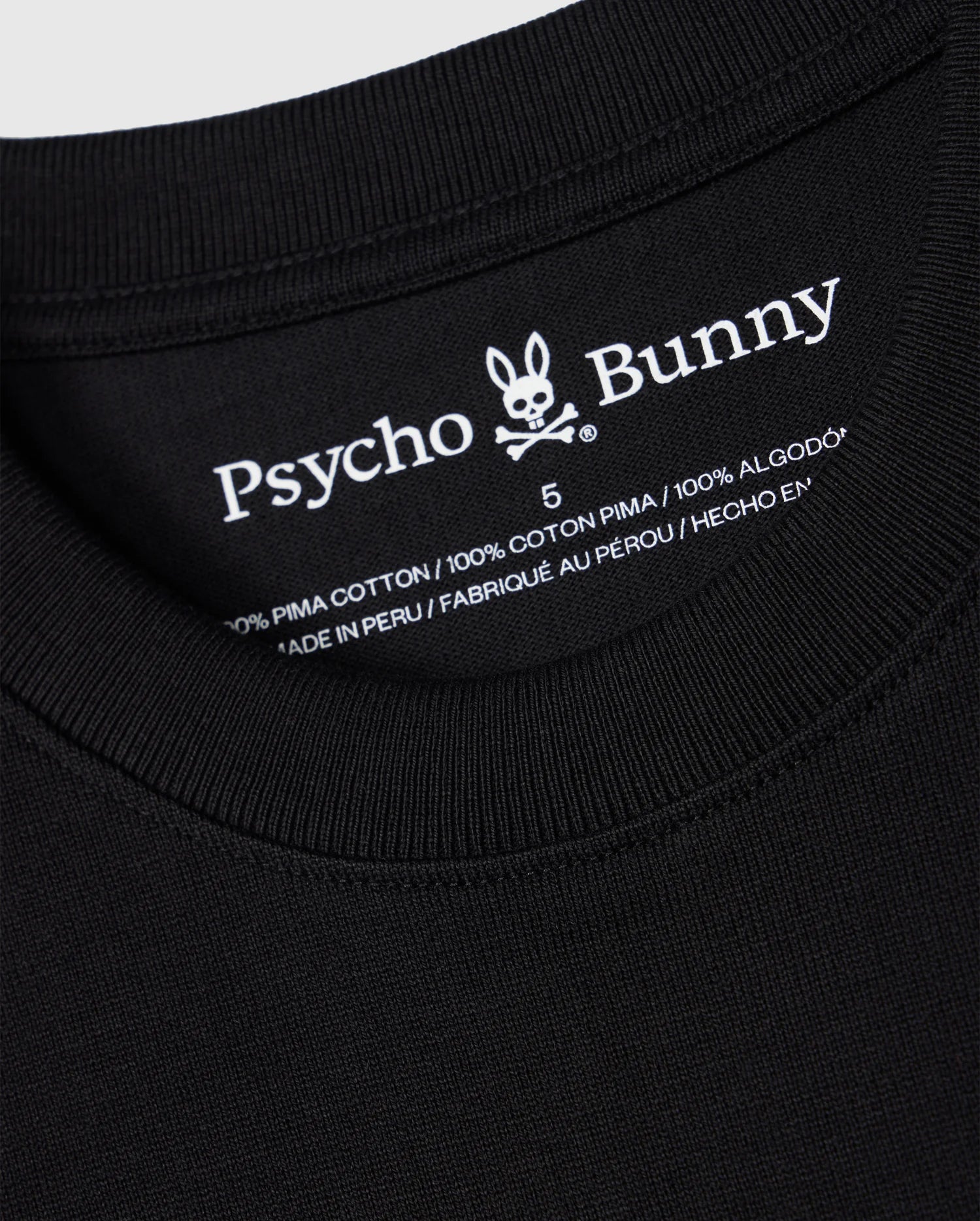 Close-up of the neckline on a black Men's Duke Oversized Tee (B6U742D200) by Psycho Bunny, showcasing a label that features the brand name and a bunny with crossbones logo. The label indicates the material as "100% Pima Cotton" and notes that this relaxed fit shirt is made in Peru.