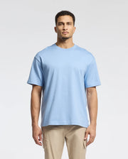A man with short hair and a neutral expression stands against a plain background wearing a Psycho Bunny MENS DUKE OVERSIZED TEE in light blue, crafted from premium Pima cotton, paired with beige pants.