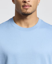 A person in a light blue Psycho Bunny MENS DUKE OVERSIZED TEE - B6U742D200, with a soft Pima cotton crew-neck and small embroidered bunny logo on the left. The relaxed fit highlights their shoulders against a plain background. They have short facial hair and an earring.
