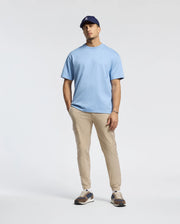 A man in a Psycho Bunny Men's Duke Oversized Tee (B6U742D200)—a blue heavyweight jersey—teams it with beige pants, a dark baseball cap, and sneakers, casually posing against a plain white background.