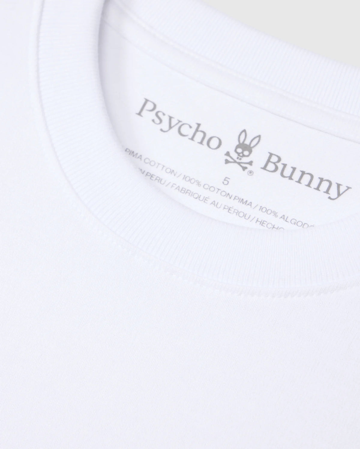Close-up of a white Psycho Bunny t-shirt label. The label displays the brand name "Psycho Bunny," a rabbit logo with crossbones, and indicates the shirt is made from 100% heavyweight Pima cotton and manufactured in Peru. The background of the image is the solid pattern fabric of this relaxed fit MENS DUKE OVERSIZED TEE - B6U742D200.
