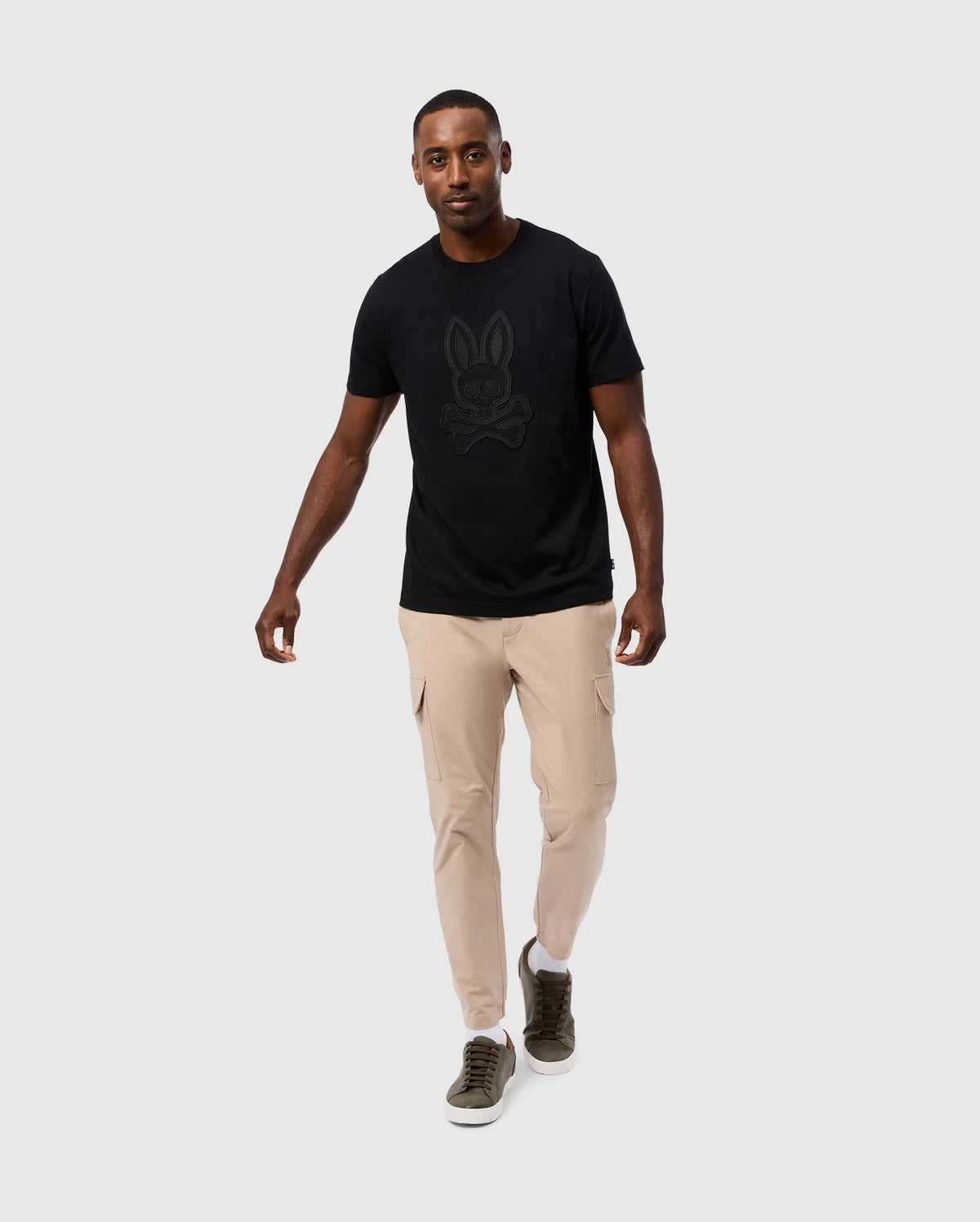 A man is walking towards the camera on a plain gray background. He is wearing a black Psycho Bunny MENS MALTON GRAPHIC TEE - B6U795D200 made from soft Pima cotton, beige cargo pants, and olive green sneakers. He has a neutral expression and one arm slightly outstretched. The high-quality t-shirt is made in Peru, adding to its unique appeal.