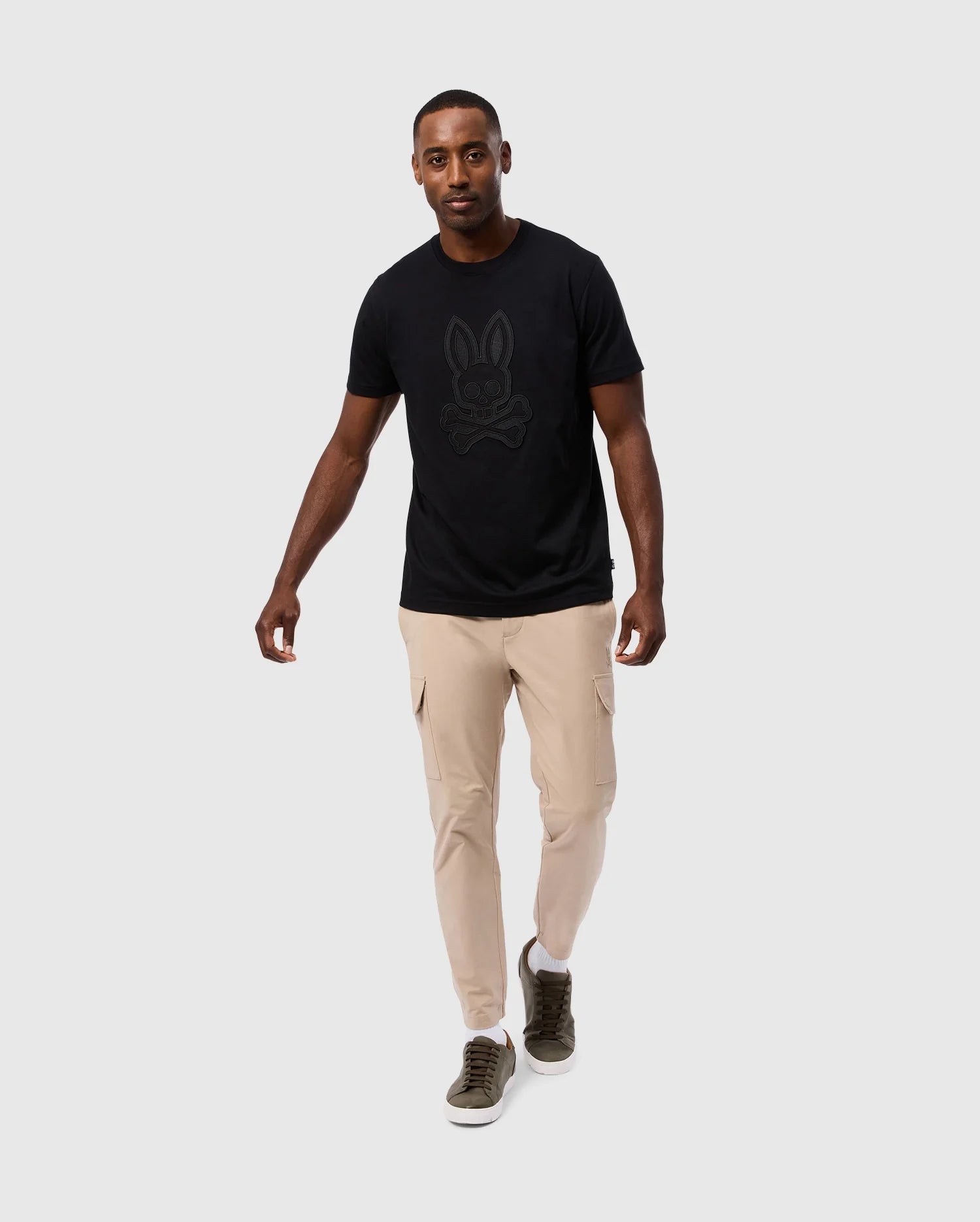 A man is walking towards the camera on a plain gray background. He is wearing a black Psycho Bunny MENS MALTON GRAPHIC TEE - B6U795D200 made from soft Pima cotton, beige cargo pants, and olive green sneakers. He has a neutral expression and one arm slightly outstretched. The high-quality t-shirt is made in Peru, adding to its unique appeal.