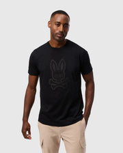 A man with a neutral expression is wearing the Psycho Bunny MENS MALTON GRAPHIC TEE - B6U795D200 made from soft Pima cotton, featuring a rabbit with crossbones design, paired with beige cargo pants. He stands against a plain, light grey background.
