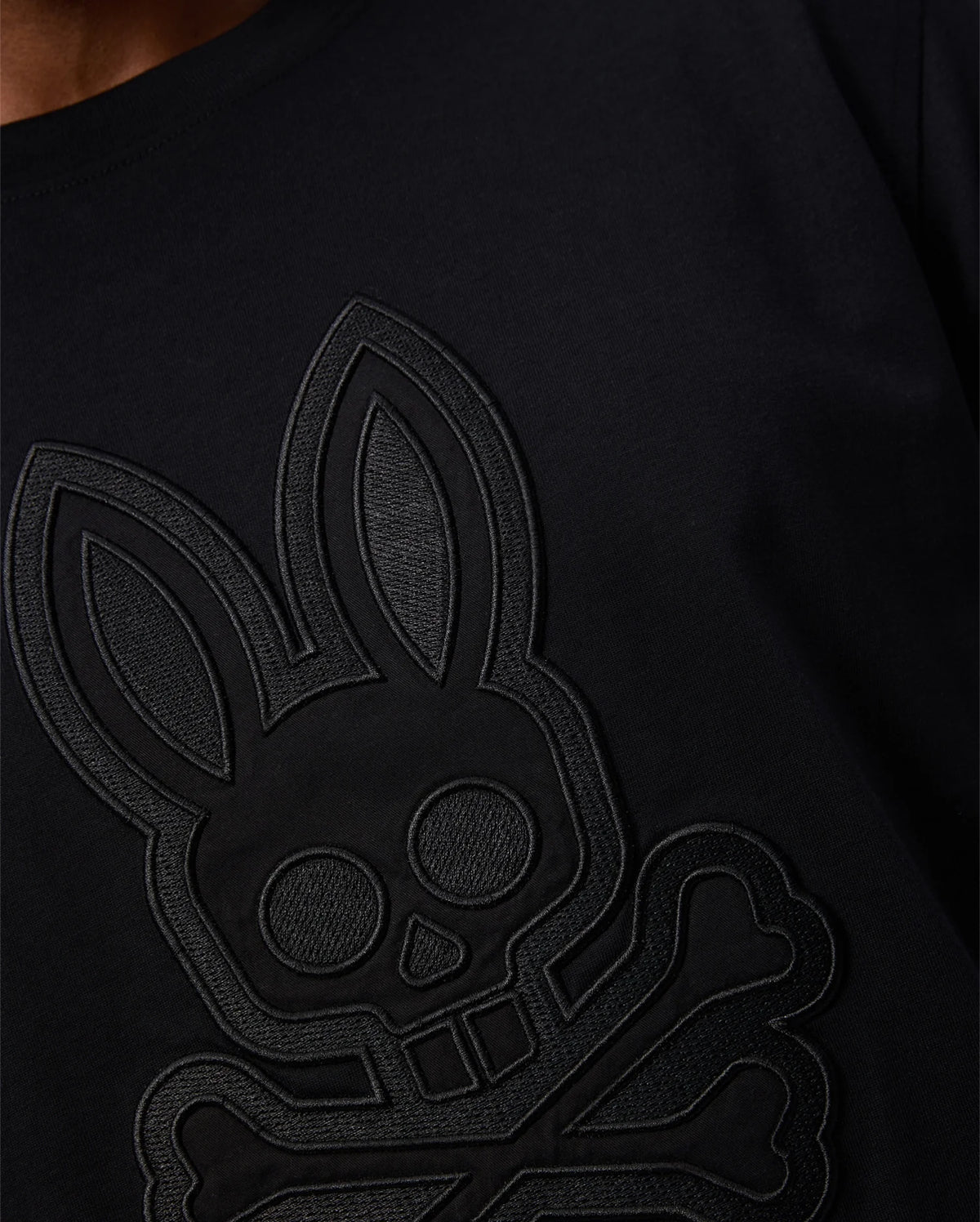 Close-up of a person wearing the Psycho Bunny MENS MALTON GRAPHIC TEE - B6U795D200 made in Peru, featuring an embossed design of a skull with bunny ears and crossbones. This Pima cotton black shirt has a textured look thanks to the raised design against the dark fabric.