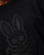 Close-up of a person wearing the Psycho Bunny MENS MALTON GRAPHIC TEE - B6U795D200 made in Peru, featuring an embossed design of a skull with bunny ears and crossbones. This Pima cotton black shirt has a textured look thanks to the raised design against the dark fabric.