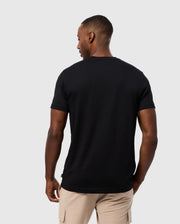 A man with short hair is standing with his back to the camera, wearing a plain black Psycho Bunny MENS MALTON GRAPHIC TEE - B6U795D200 made of soft Pima cotton, paired with beige cargo shorts. The background is a solid light color.