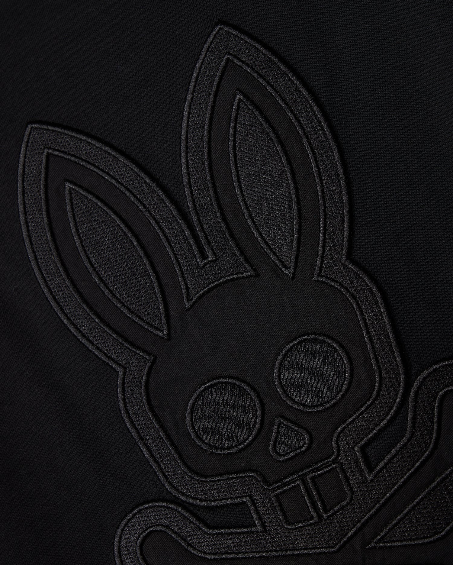 A close-up view of the black-on-black embroidered patch on the Psycho Bunny MENS MALTON GRAPHIC TEE - B6U795D200 reveals a stylized bunny skull and crossbones design. The subtle, raised outlines and details on the Pima cotton fabric create a minimalistic and edgy look. Crafted in Peru for exceptional quality.